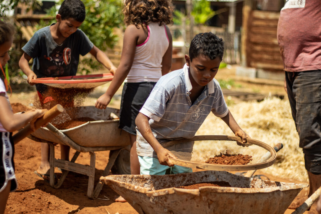 Crianças peneiram terra no canteiro, com carrinhos de mão